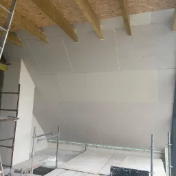 Montaż płyt gipsowo-kartonowych na poddaszu, widoczne drewniane belki stropowe, aluminiowa drabina i rusztowanie, okno w ciemnej ramie.