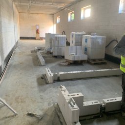 BARVINOVA BUD - Wnętrze w trakcie murowania ścian z białych bloczków. Na podłodze leżą narzędzia i ułożone fragmenty muru. W tle widoczne palety z zapakowanymi bloczkami.