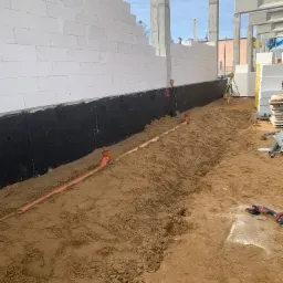 Fundamenty budynku z białych bloczków, zabezpieczone czarną izolacją. W wykopie ułożone rury kanalizacyjne. Widoczny sprzęt budowlany i ziemia.