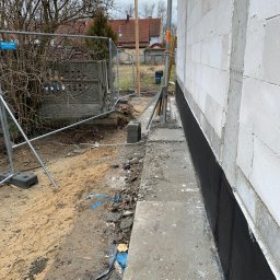 BARVINOVA BUD - Ściana budynku z białych bloczków, zabezpieczona czarną izolacją przeciwwilgociową. Widoczne fundamenty i teren budowy z ogrodzeniem tymczasowym w tle.