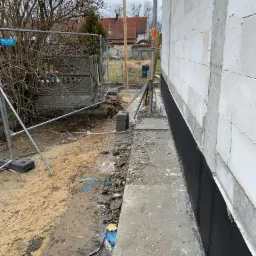 Ściana budynku z białych bloczków, zabezpieczona czarną izolacją przeciwwilgociową. Widoczne fundamenty i teren budowy z ogrodzeniem tymczasowym w tle.