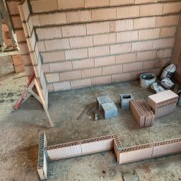 BARVINOVA BUD - Wnętrze w trakcie murowania ścian z bloczków ceramicznych, widoczne narzędzia i materiały budowlane. Fragment ściany już wzniesiony, w tle worki z zaprawą.