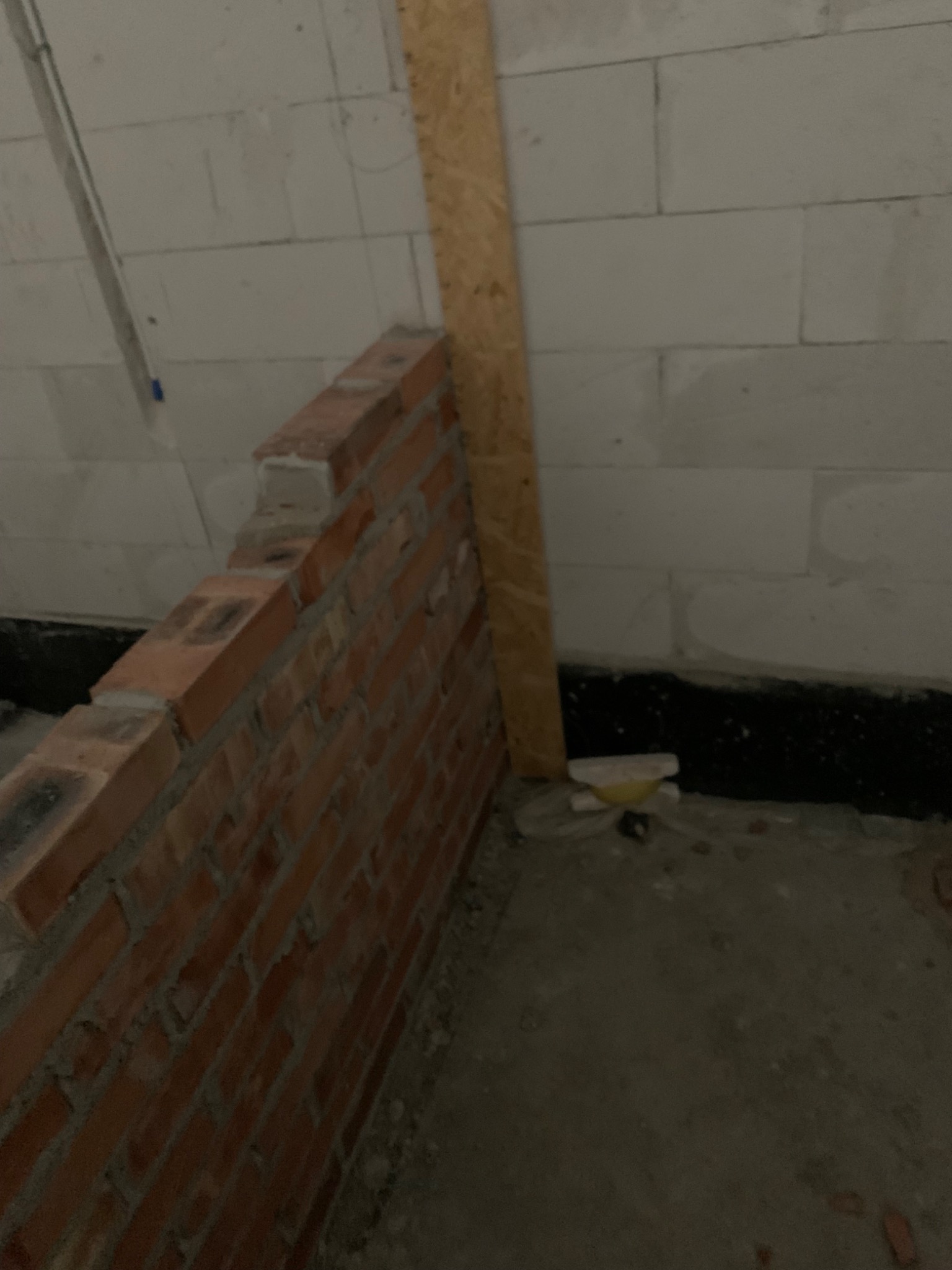 Częściowo wymurowana ściana z czerwonej cegły, oparta o drewnianą płytę, na tle ściany z białych bloczków. Widoczny fragment posadzki i instalacji.