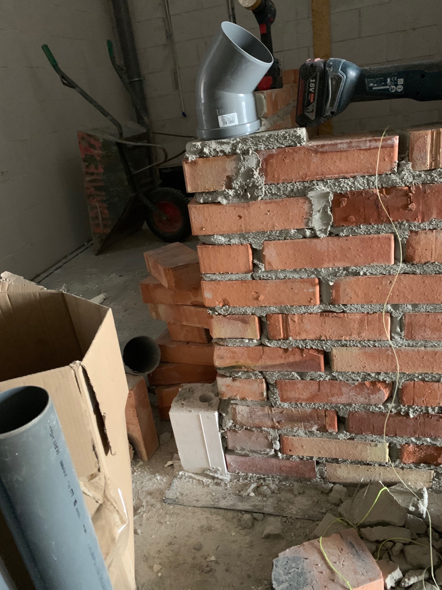 Murowana ściana z czerwonej cegły w trakcie budowy, widoczne narzędzia i materiały budowlane, w tym rura wentylacyjna i piła akumulatorowa.