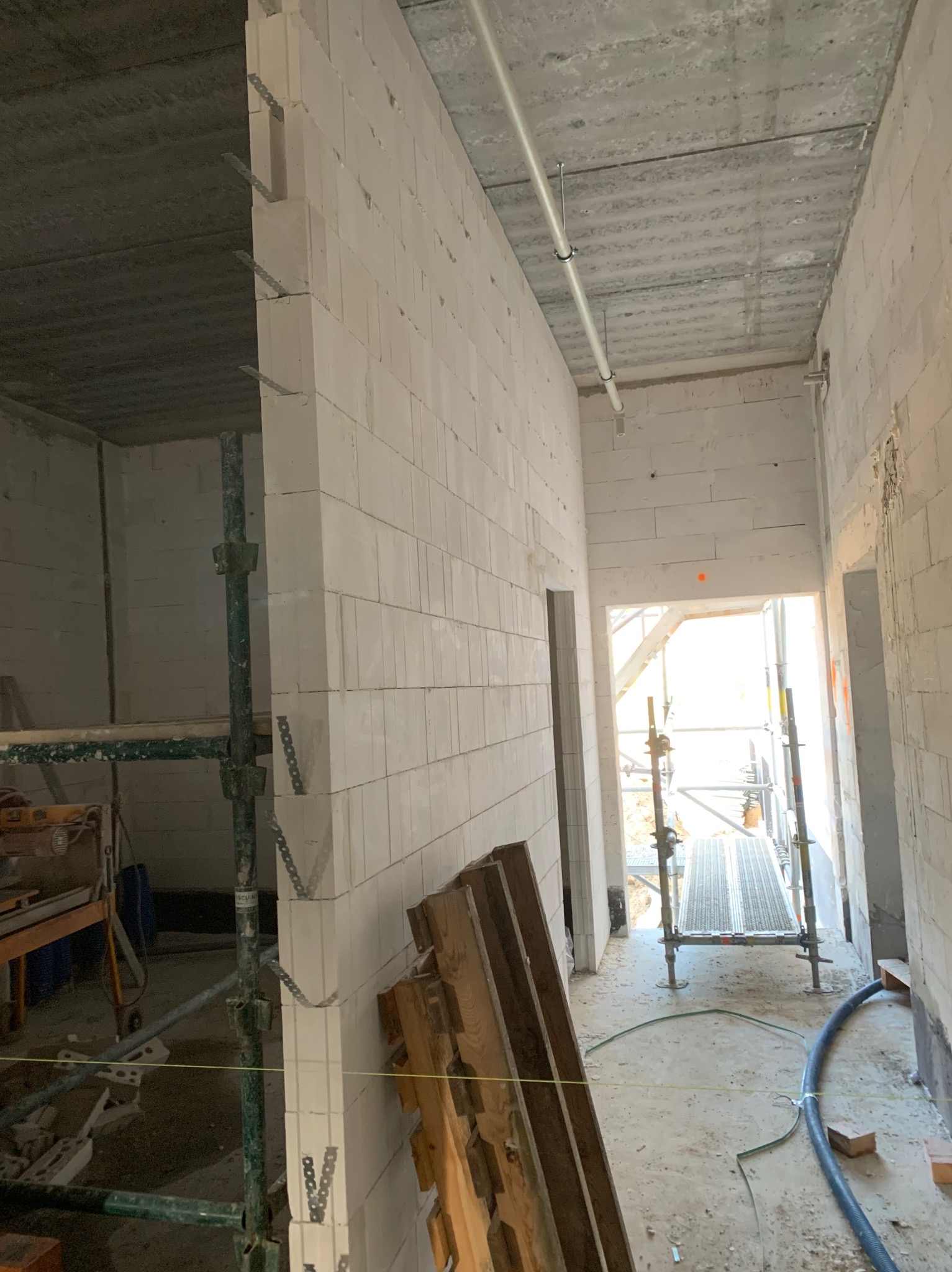 Wnętrze w trakcie budowy: białe ściany z betonu komórkowego, widoczne rusztowania i narzędzia. W oddali otwór drzwiowy z widokiem na zewnątrz.