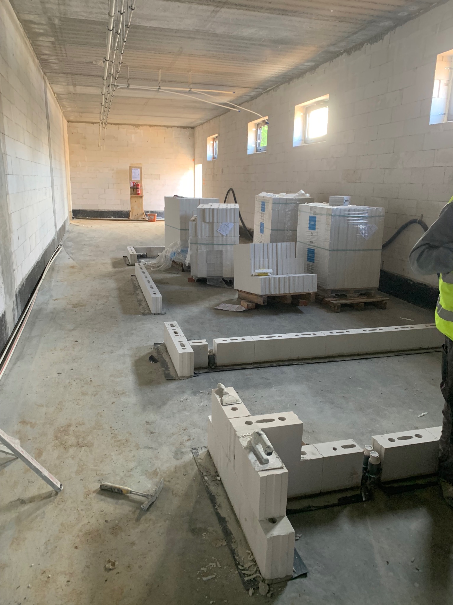 Wnętrze w trakcie murowania ścian z białych bloczków. Na podłodze leżą narzędzia i ułożone fragmenty muru. W tle widoczne palety z zapakowanymi bloczkami.