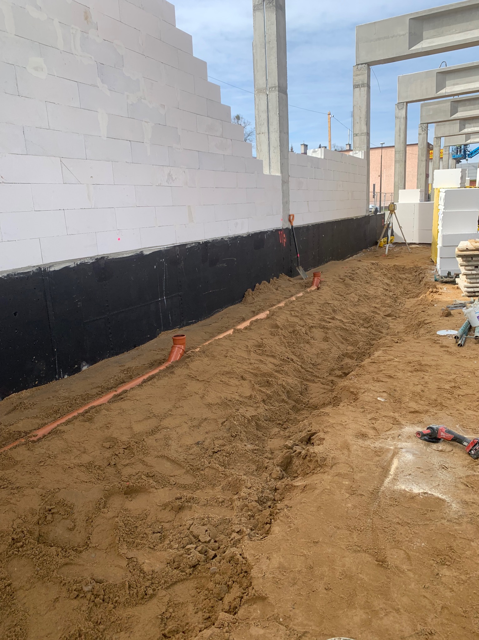 Fundamenty budynku z białych bloczków, zabezpieczone czarną izolacją. W wykopie ułożone rury kanalizacyjne. Widoczny sprzęt budowlany i ziemia.