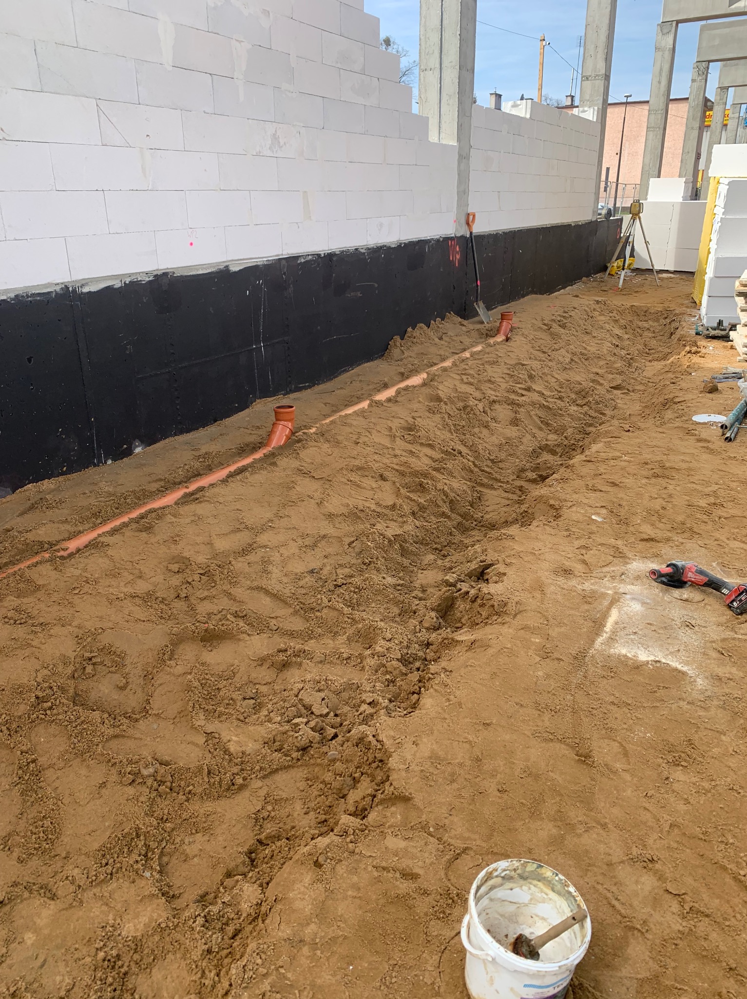 Fundamenty domu w trakcie budowy: widoczne rury kanalizacyjne w wykopie, ściany z białych bloczków i sprzęt pomiarowy. Etap prac ziemnych i instalacyjnych.