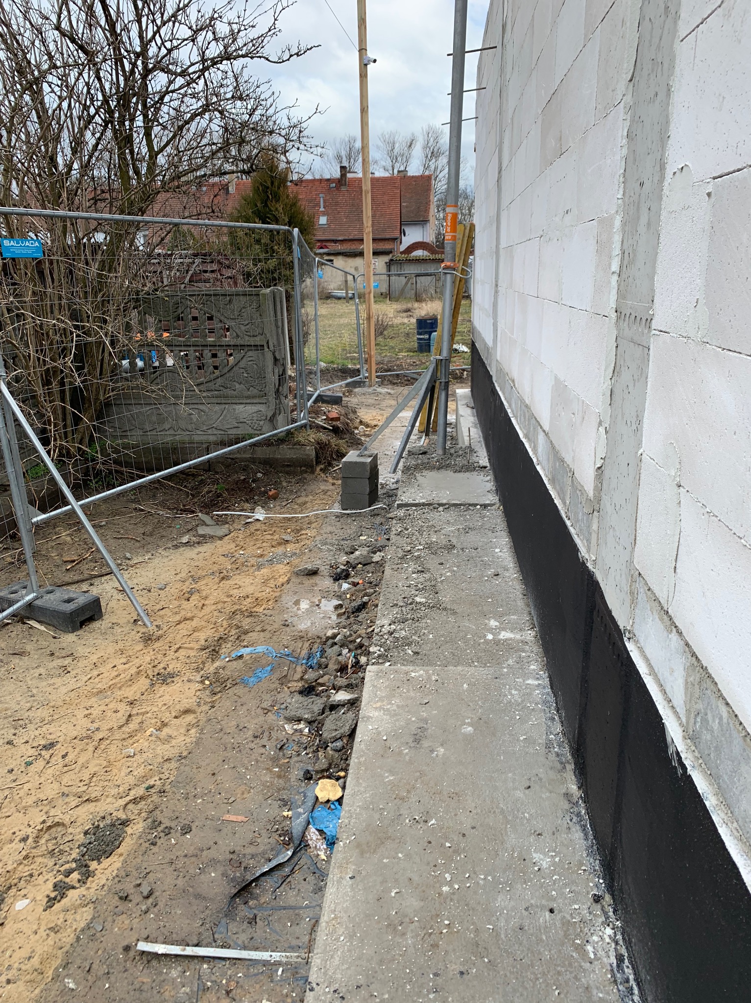 Ściana budynku z białych bloczków, zabezpieczona czarną izolacją przeciwwilgociową. Widoczne fundamenty i teren budowy z ogrodzeniem tymczasowym w tle.