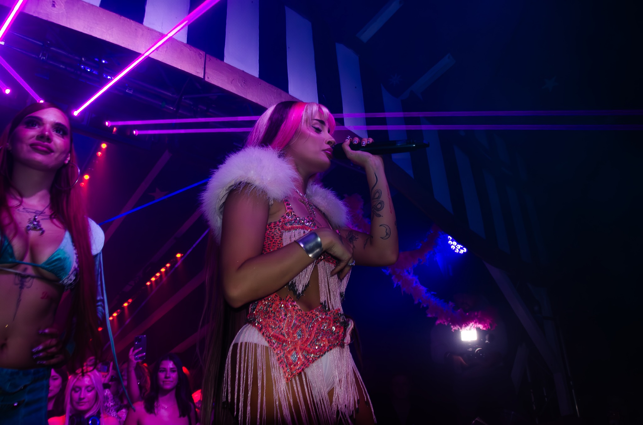 Koncertowe ujęcie artystki z różowymi włosami, w ozdobnym stroju z frędzlami, śpiewającej do mikrofonu na scenie w klubie z laserami i publicznością.