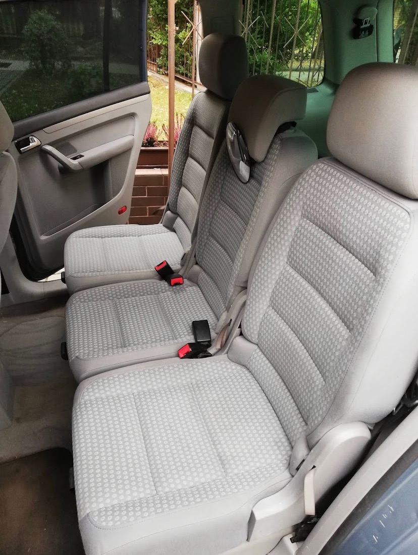 Wnętrze samochodu: tylne siedzenia z jasnoszarą tapicerką w drobny wzór, widoczne pasy bezpieczeństwa i zagłówki. Czyste i zadbane.