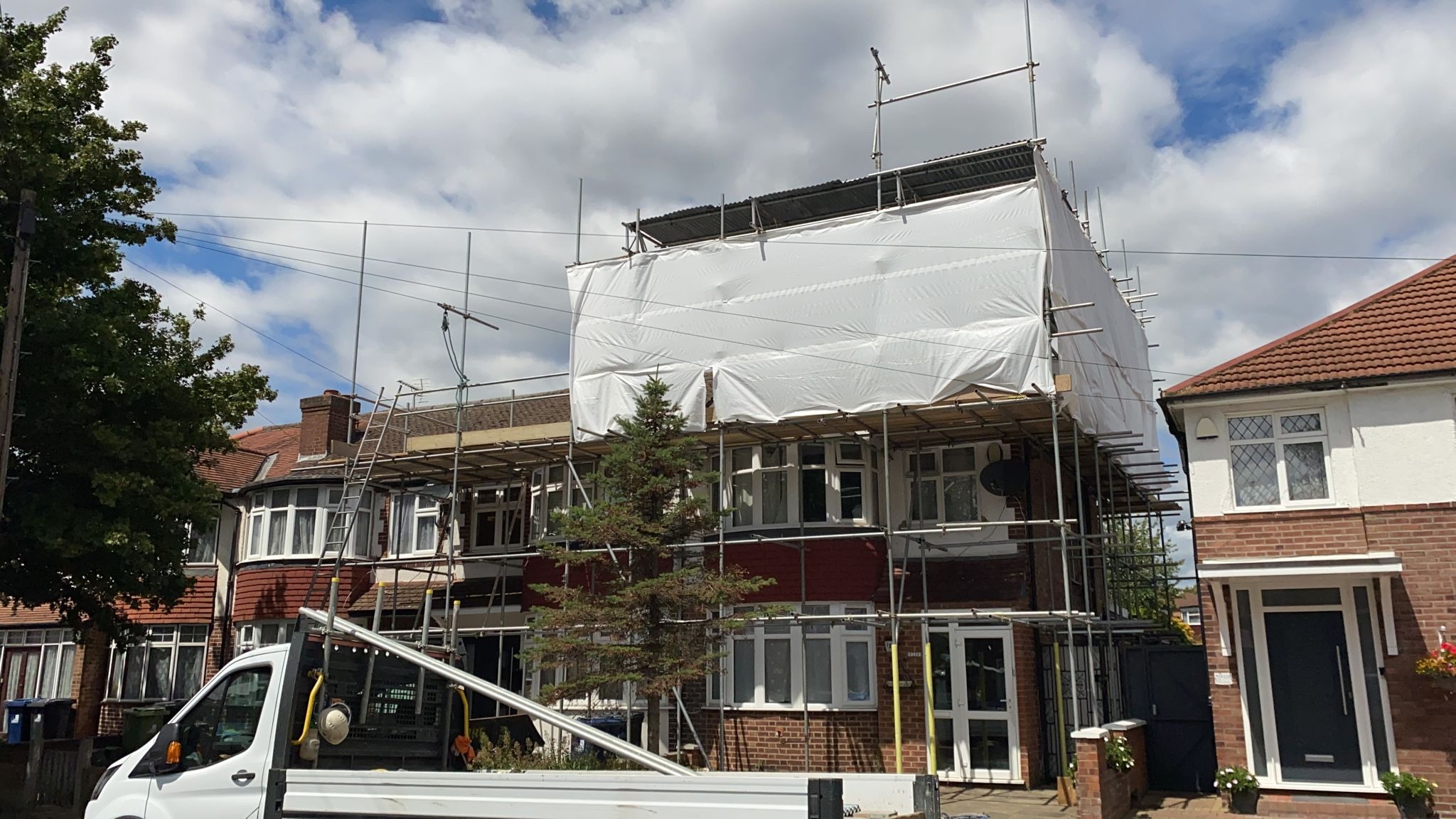 Dwupiętrowy dom w rusztowaniach, osłonięty białą plandeką, z widocznym fragmentem białej furgonetki na pierwszym planie i drzewem iglastym zasłaniającym część fasady.