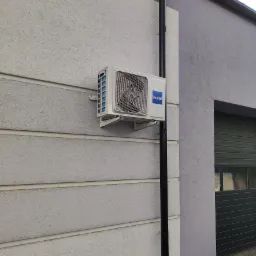 Zewnętrzna jednostka klimatyzacji marki Haier zamontowana na ścianie budynku z szarą elewacją, obok rury spustowej.