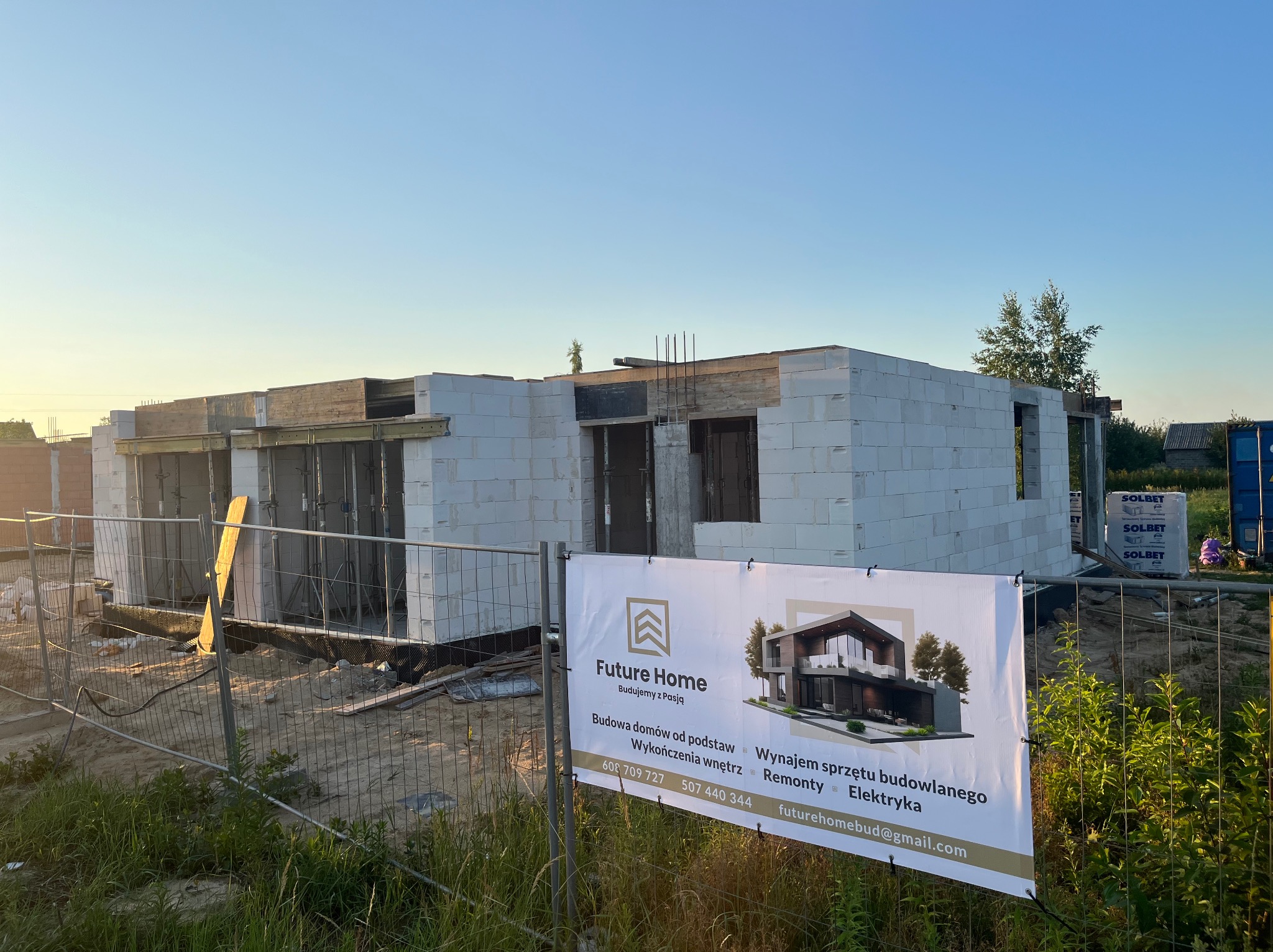 Wczesny etap budowy domu z białych bloczków, widoczne zbrojenia i szalunki, ogrodzenie budowy z banerem reklamowym firmy budowlanej Future Home.
