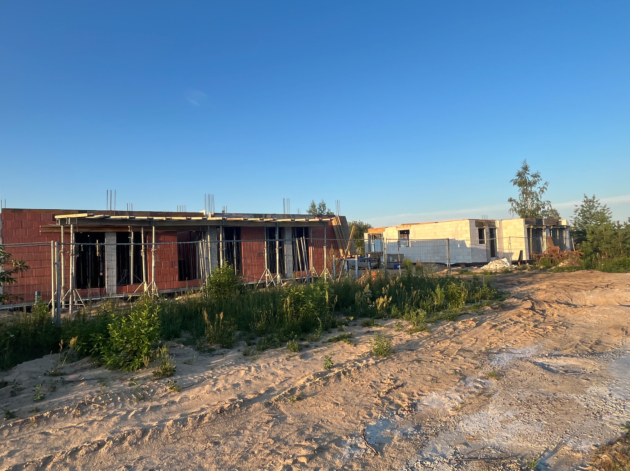 Dwa budynki w stanie surowym otwartym z czerwonej cegły i białego gazobetonu, widoczne z poziomu gruntu, na pierwszym planie piasek i roślinność, zabezpieczone ogrodzeniem tymczasowym.
