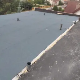 Płaski dach pokryty szarą papą termozgrzewalną z wystającymi kominkami wentylacyjnymi, widoczne obróbki blacharskie i narzędzia pracy, w tle zabudowa jednorodzinna.