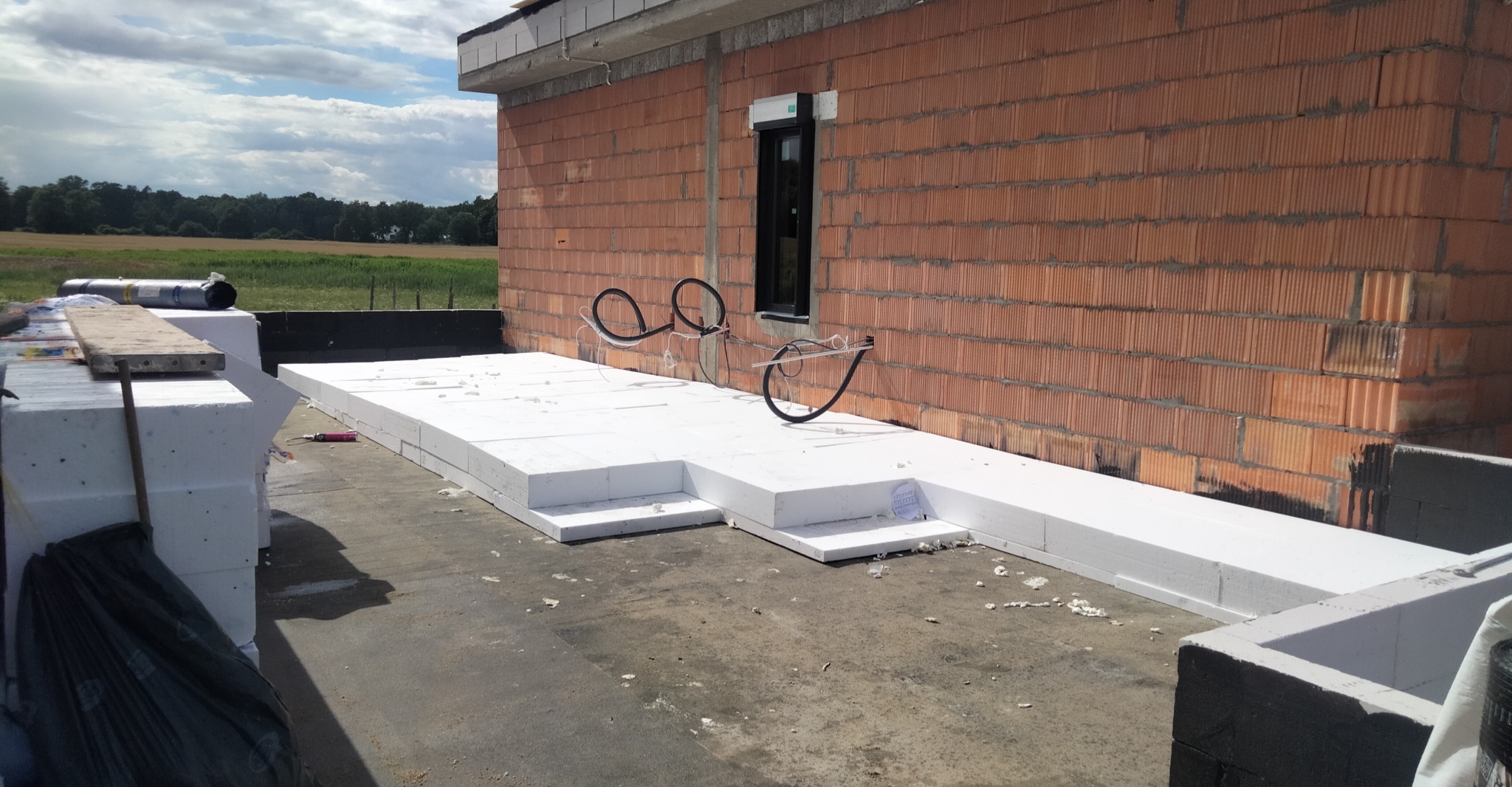 Budowa domu: ściana z czerwonej cegły, okno z roletą zewnętrzną i wystające kable elektryczne. Na dachu ułożone płyty styropianowe.