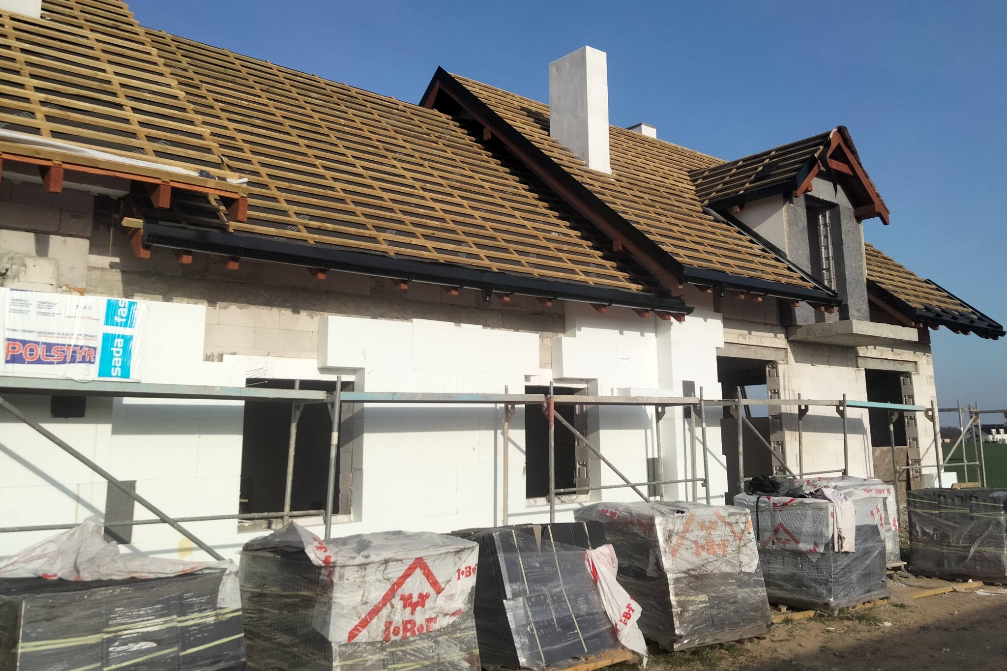 Budowa domu w trakcie realizacji: wzniesione ściany z białego bloczka, ocieplenie styropianem, przygotowany ruszt pod pokrycie dachowe, widoczne kominy i palety z materiałami budowlanymi. Ujęcie...