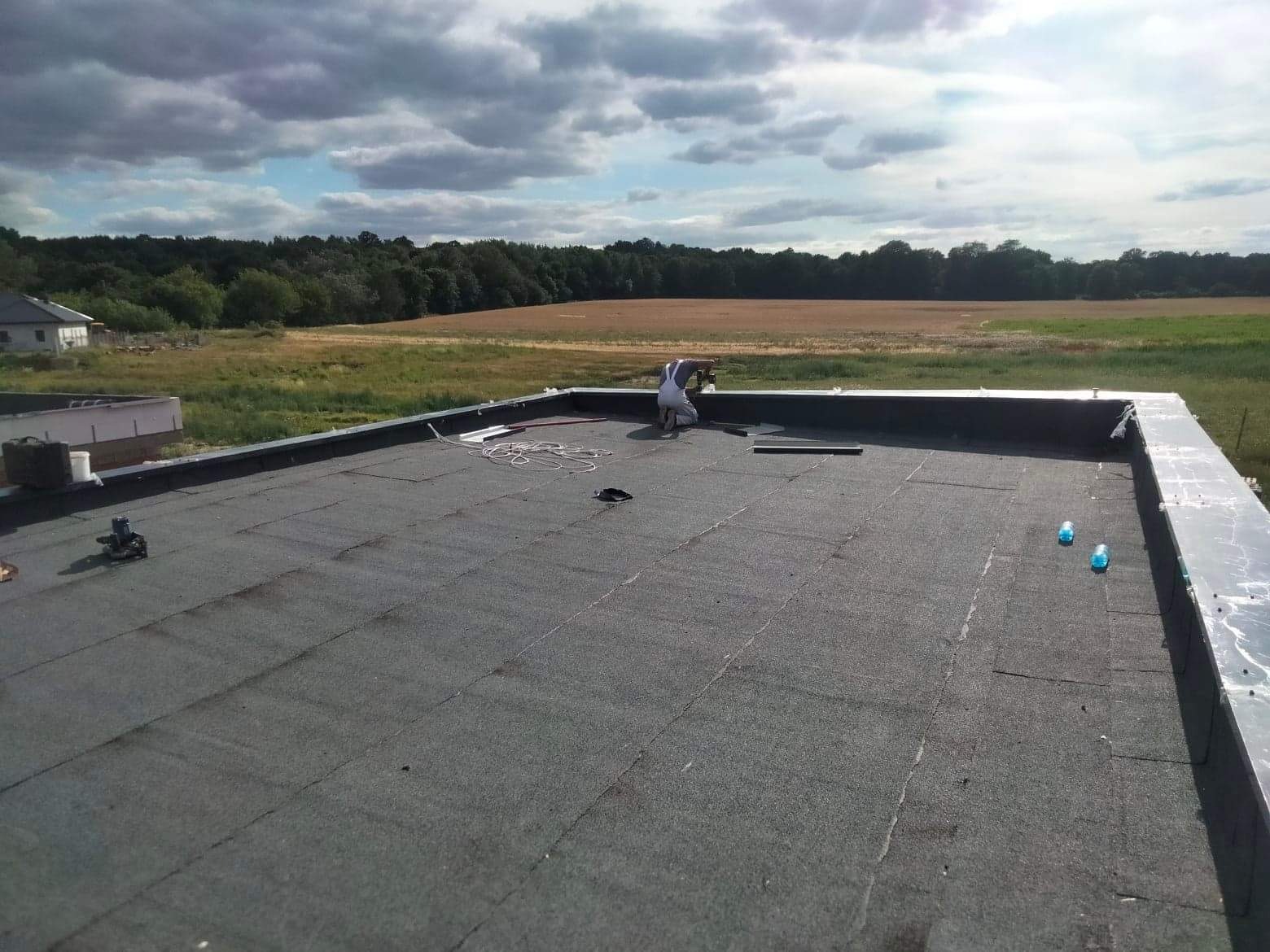 Pracownik w białym kombinezonie wykonuje uszczelnianie płaskiego dachu pokrytego papą na tle zieleni i pochmurnego nieba.
