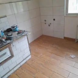 Wnętrze łazienki w trakcie układania drewnopodobnych płytek podłogowych, widoczne krzyżyki dystansowe i narzędzia na blacie obłożonym białymi kaflami.