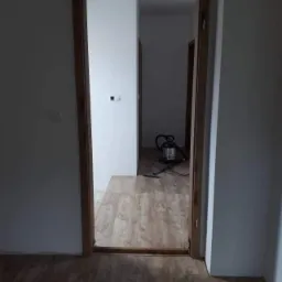 Widok na świeżo położoną podłogę laminowaną w odcieniach drewna, widoczną przez drewnianą ościeżnicę drzwiową. W tle, odkurzacz przemysłowy na podłodze w kolejnym pomieszczeniu.