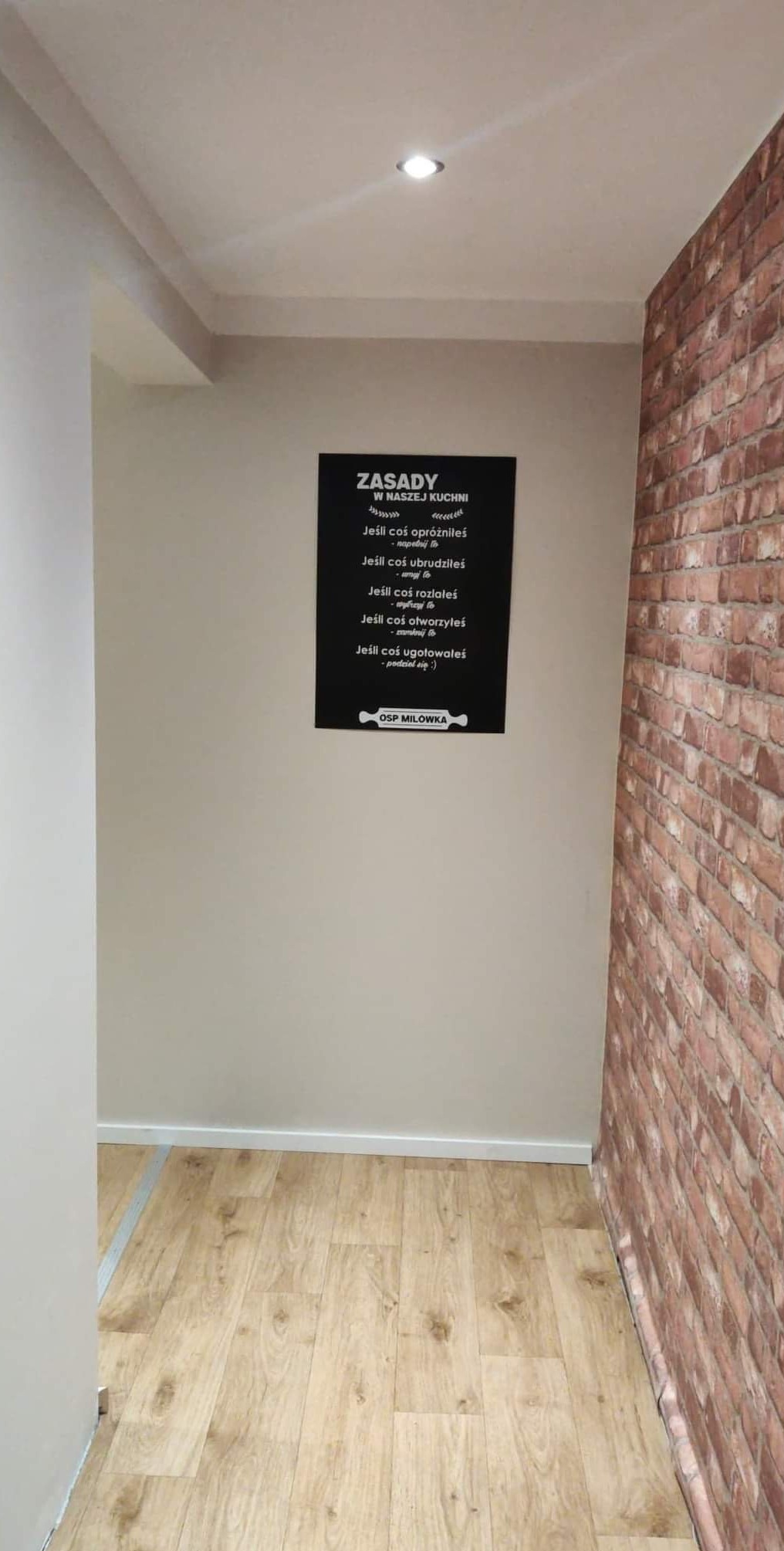 Korytarz z drewnopodobną podłogą i ścianą z cegły, na ścianie wisi czarna tablica z białymi napisami 'Zasady w naszej kuchni' oraz logo OSP Milówka.
