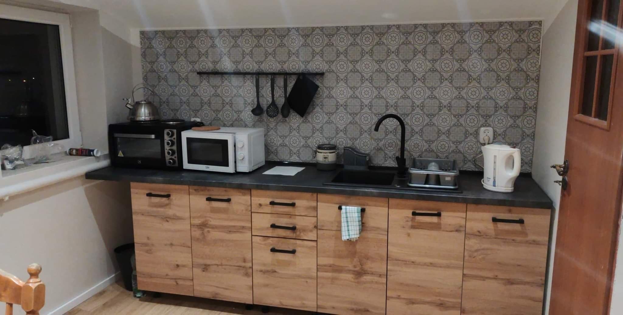 Nowoczesna kuchnia z drewnianymi szafkami, czarnym blatem i ozdobną tapetą w geometryczne wzory, wyposażona w piekarnik, mikrofalówkę i czarny zlew z baterią.