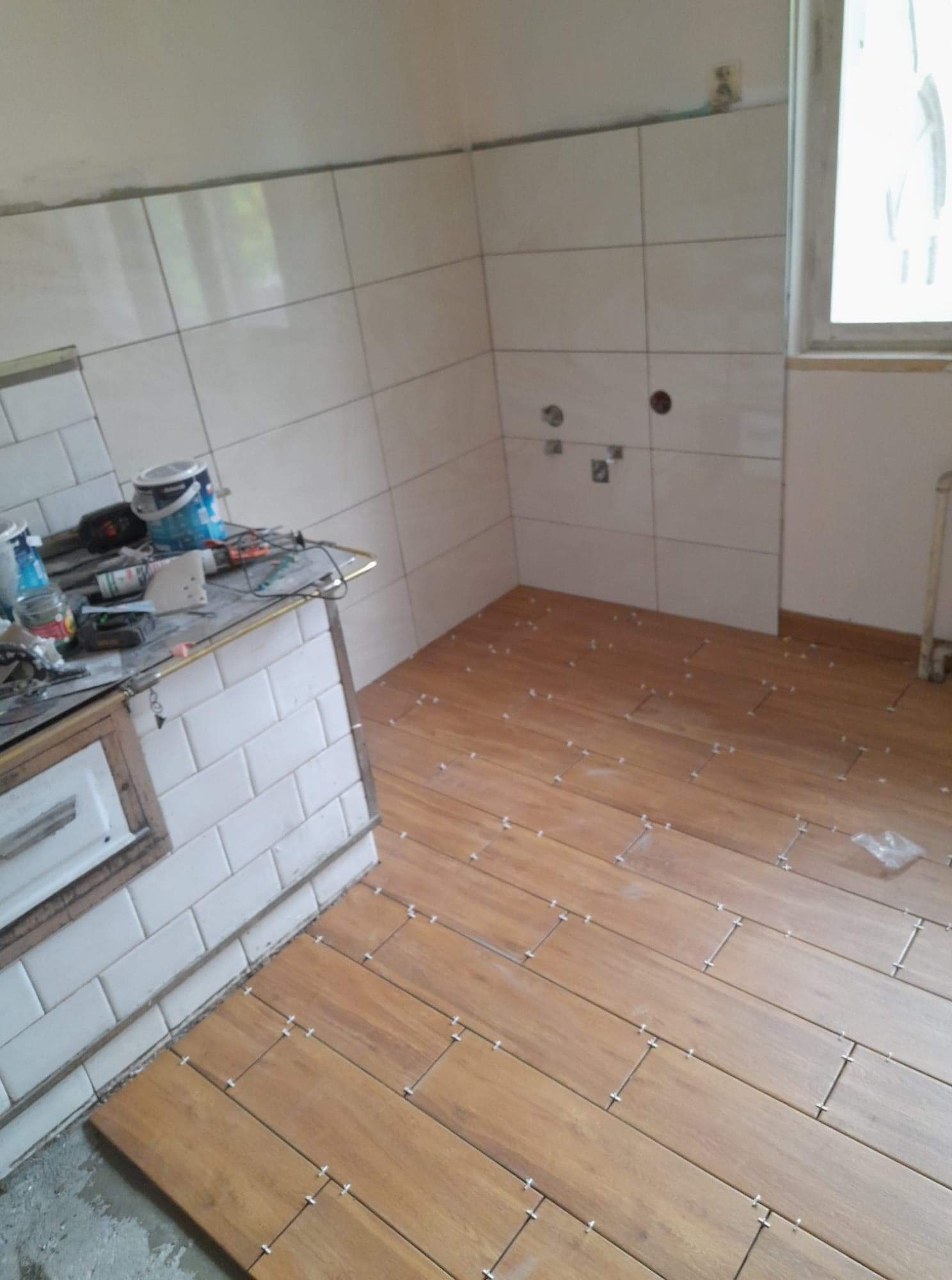 Wnętrze łazienki w trakcie układania drewnopodobnych płytek podłogowych, widoczne krzyżyki dystansowe i narzędzia na blacie obłożonym białymi kaflami.