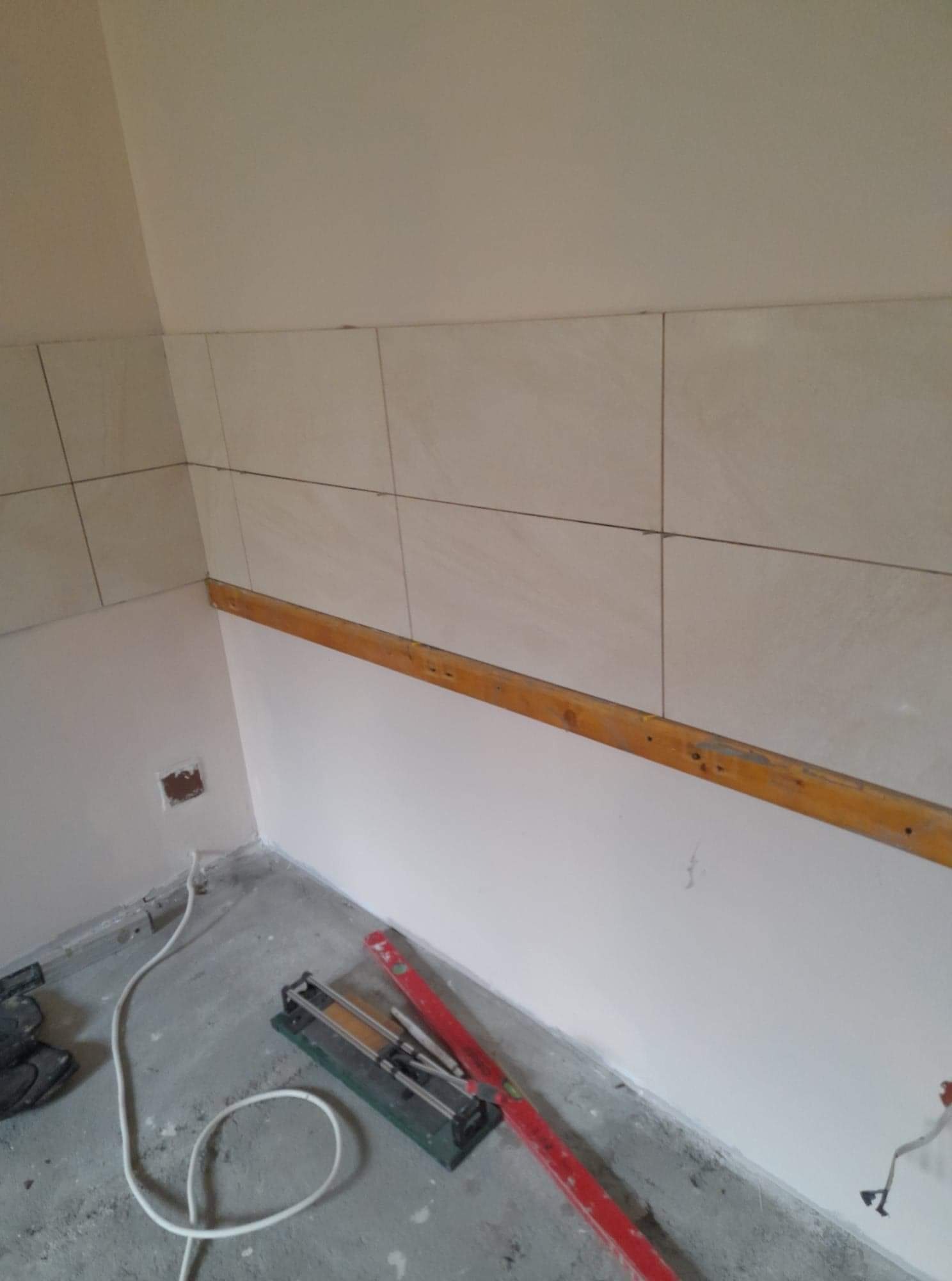 Fragment ściany z częściowo ułożonymi beżowymi płytkami, drewniana listwa poziomująca, przecinarka do płytek oraz poziomica na podłodze w trakcie prac wykończeniowych.