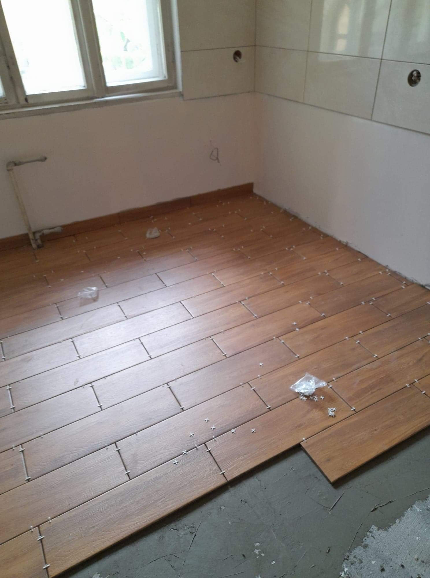 Układanie podłogowych płytek imitujących drewno w pomieszczeniu z oknem i fragmentem wykafelkowanej ściany; widoczne plastikowe krzyżyki dystansowe i otwarte opakowanie z nimi.