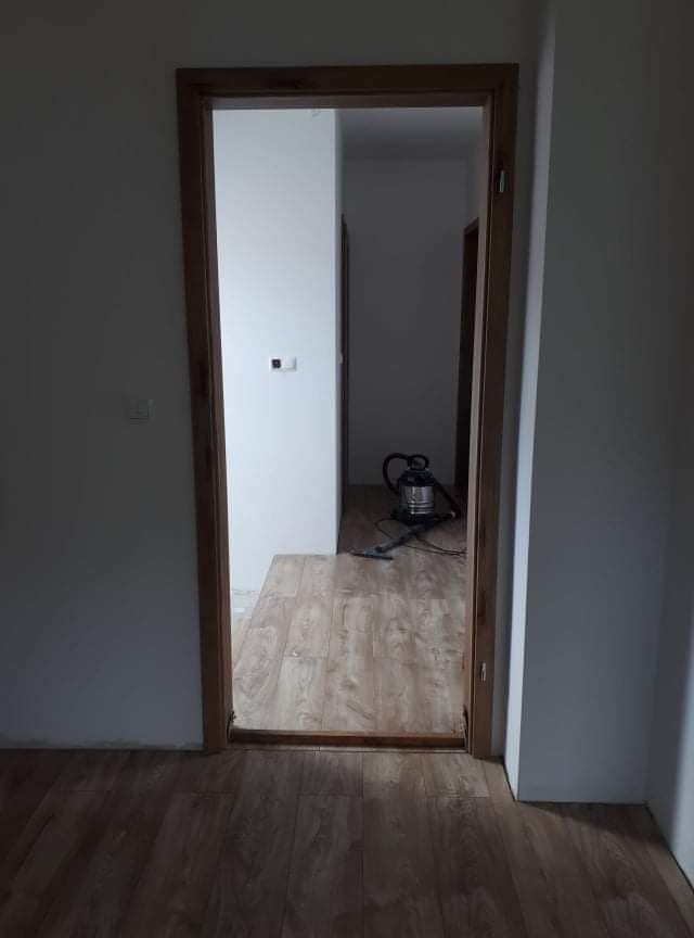 Widok na świeżo położoną podłogę laminowaną w odcieniach drewna, widoczną przez drewnianą ościeżnicę drzwiową. W tle, odkurzacz przemysłowy na podłodze w kolejnym pomieszczeniu.