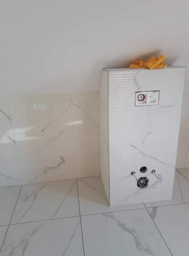 Stelaż podtynkowy obudowany płytkami imitującymi marmur, z widocznymi otworami na rury i przyciskiem spłukującym, w łazience z białymi płytkami z szarymi żyłkami, pomarańczowa szmatka na górze.