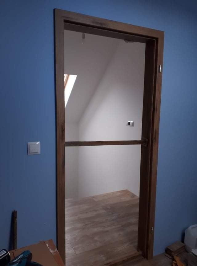 Nowa drewniana ościeżnica z zamontowanymi drzwiami w surowym stanie, widoczna na tle niebieskiej ściany i poddasza z oknem dachowym, na podłodze panele imitujące drewno.