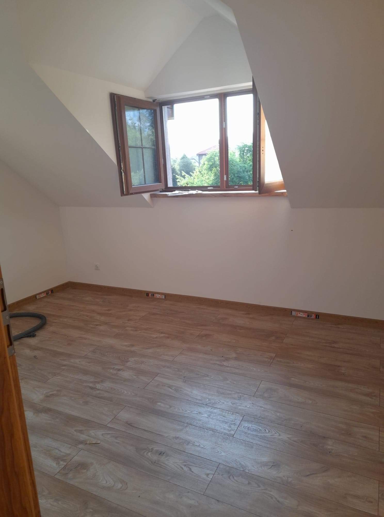 Poddasze po remoncie z drewnianą podłogą, otwartym oknem z brązową ramą i białymi ścianami, widoczny odkurzacz i fragment drzwi.