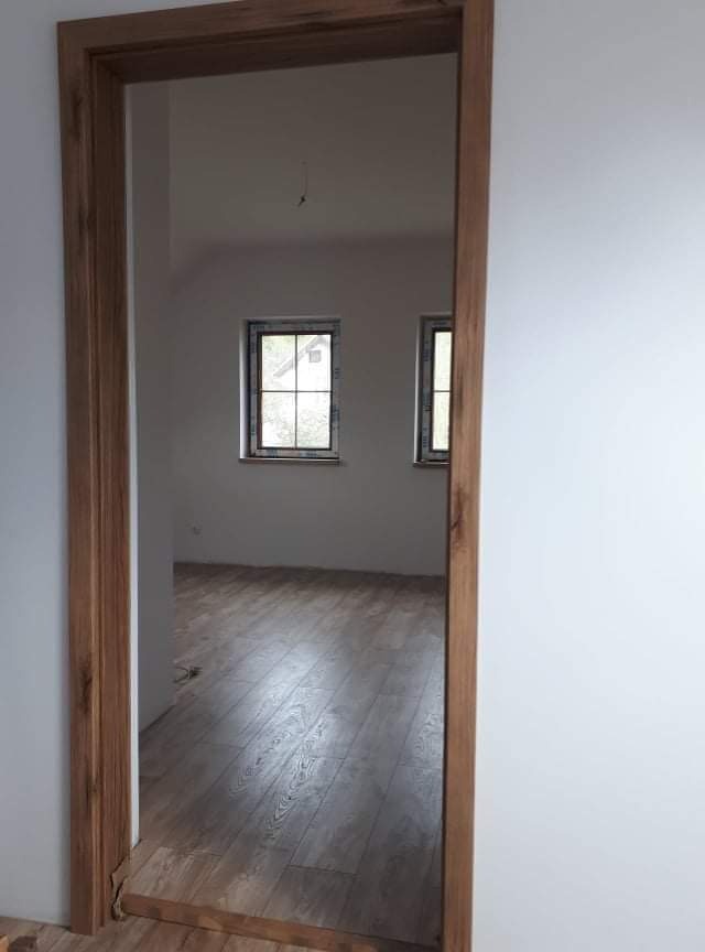 Drewniana ościeżnica w nowo wykończonym pokoju z panelami podłogowymi i widocznymi dwoma oknami, ściany pomalowane na biało, widoczne niedokończone wykończenie podłogi przy ościeżnicy.