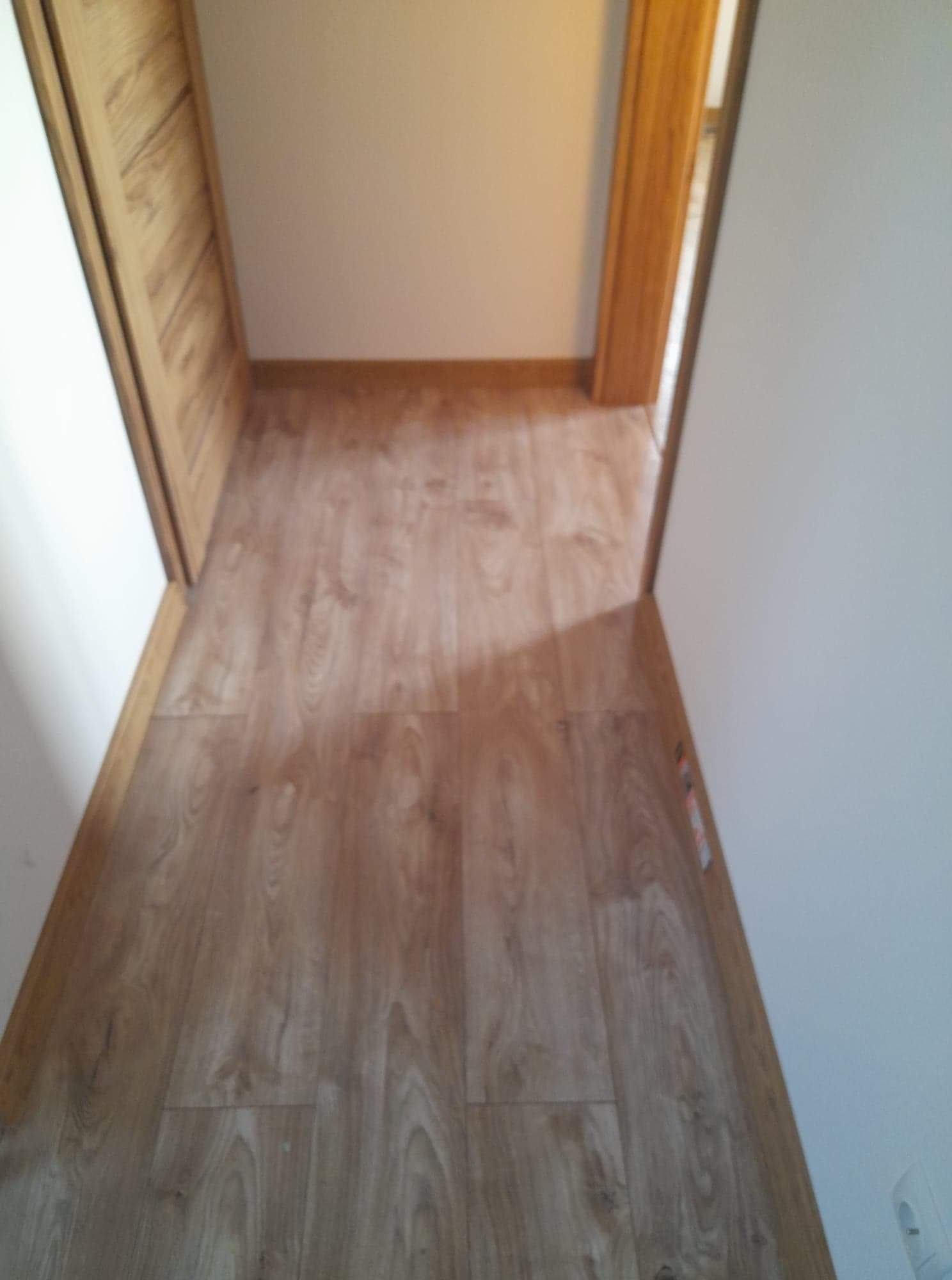 Ułożona podłoga laminowana w wąskim korytarzu z białymi ścianami i drewnianymi ościeżnicami drzwi, widziana z góry, z widocznymi łączeniami paneli i listwami przypodłogowymi.