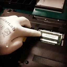 Otwarty laptop z widoczną pamięcią RAM, instalowaną przez osobę w białej rękawiczce ochronnej SolderLab EN388. Widoczny fragment obudowy i tacka na napęd.