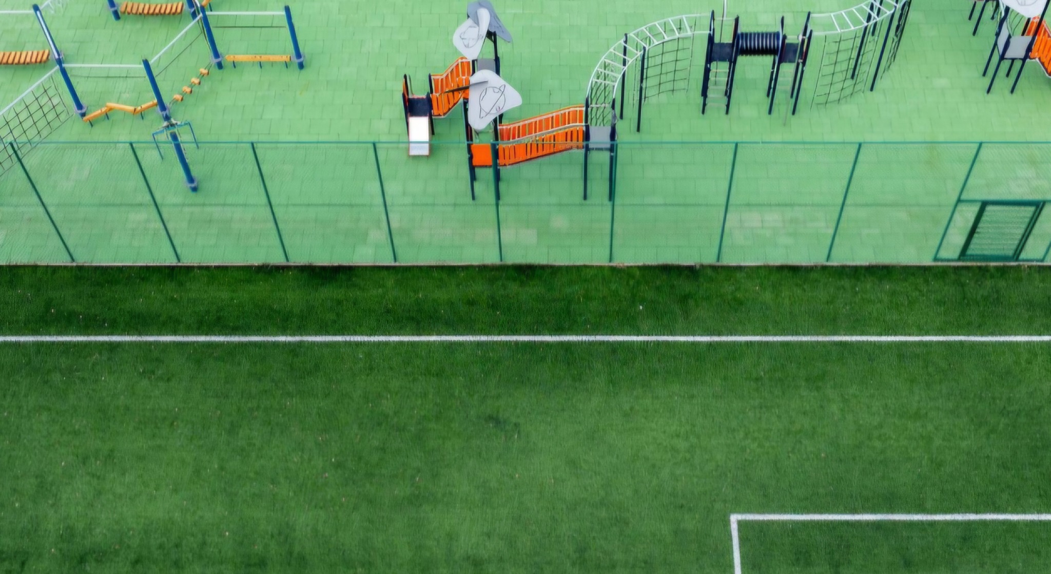 Widok z góry na plac zabaw z zieloną nawierzchnią i różnorodnymi urządzeniami do zabawy, oddzielony od boiska sportowego zielonym ogrodzeniem.