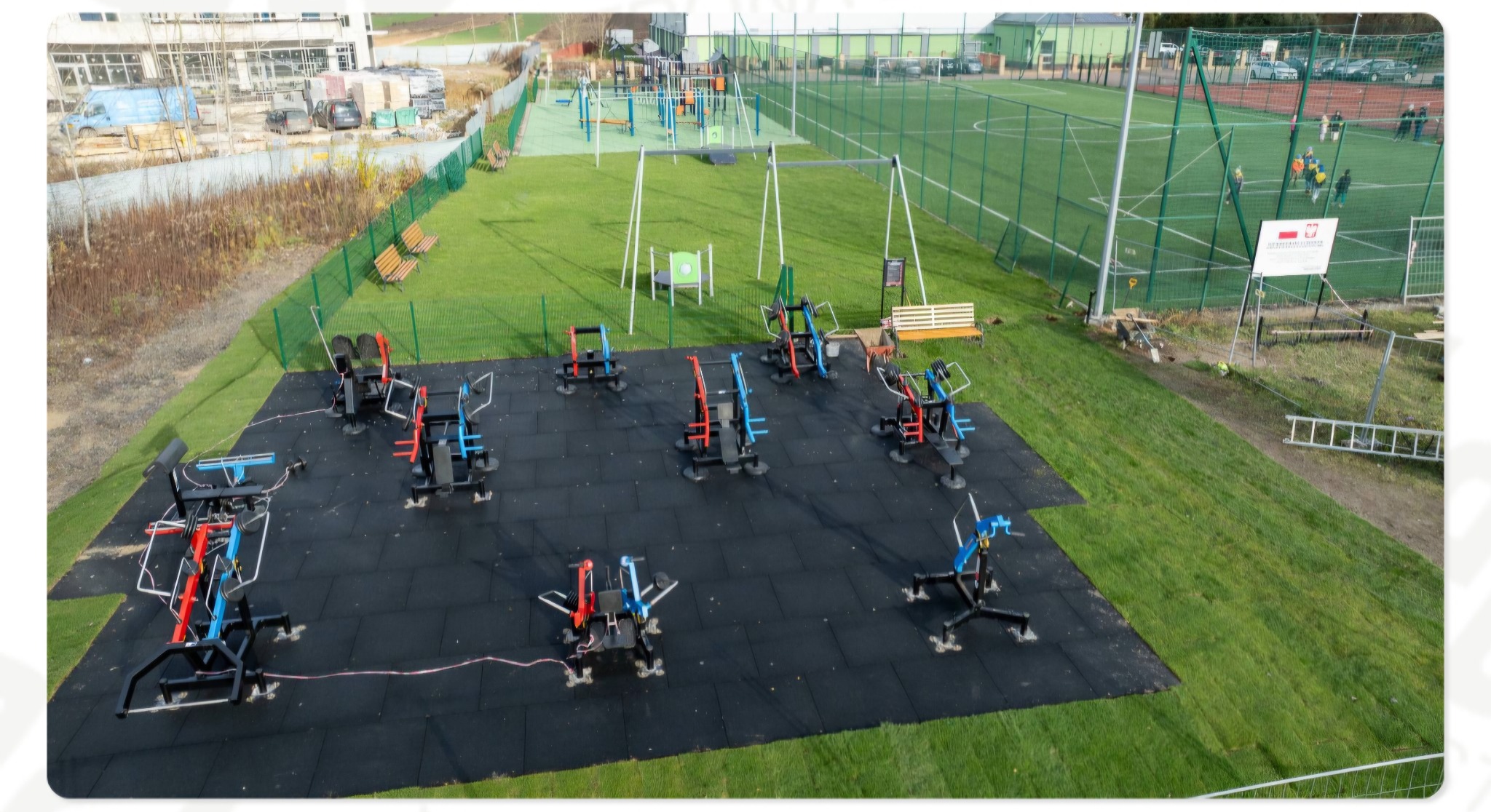 Widok z góry na nowo wybudowaną, zewnętrzną siłownię z czarną, gumową nawierzchnią i kolorowymi urządzeniami do ćwiczeń na tle boiska i placu zabaw.