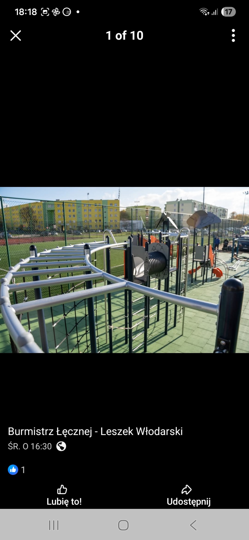 Nowoczesny plac zabaw z metalowymi konstrukcjami do wspinaczki i ćwiczeń, na tle zielonego boiska i budynków mieszkalnych w słoneczny dzień.