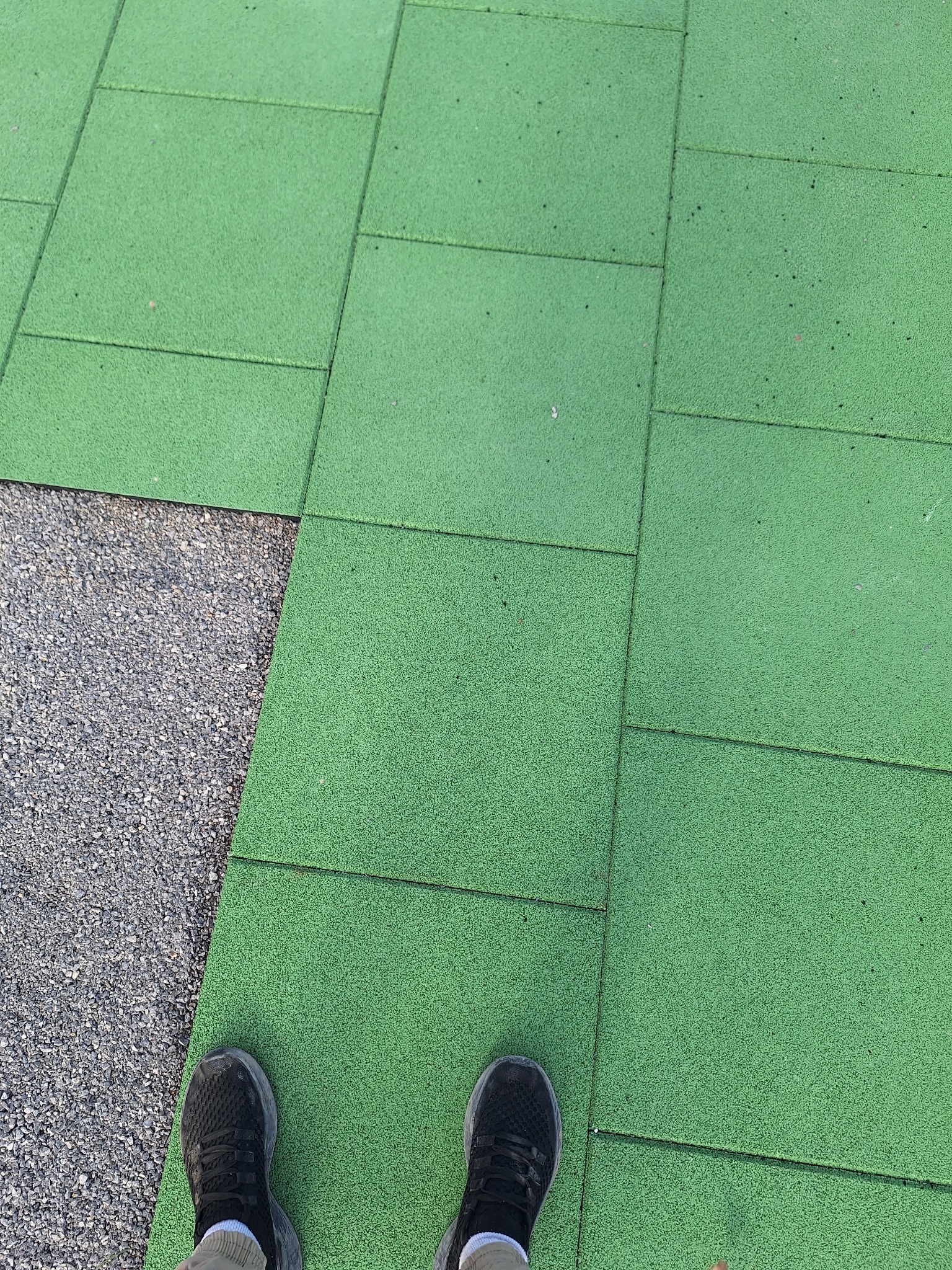 Zielona, gumowa nawierzchnia placu zabaw ułożona z kwadratowych płyt, widoczne buty osoby stojącej na nawierzchni, obok szara, żwirowa powierzchnia.