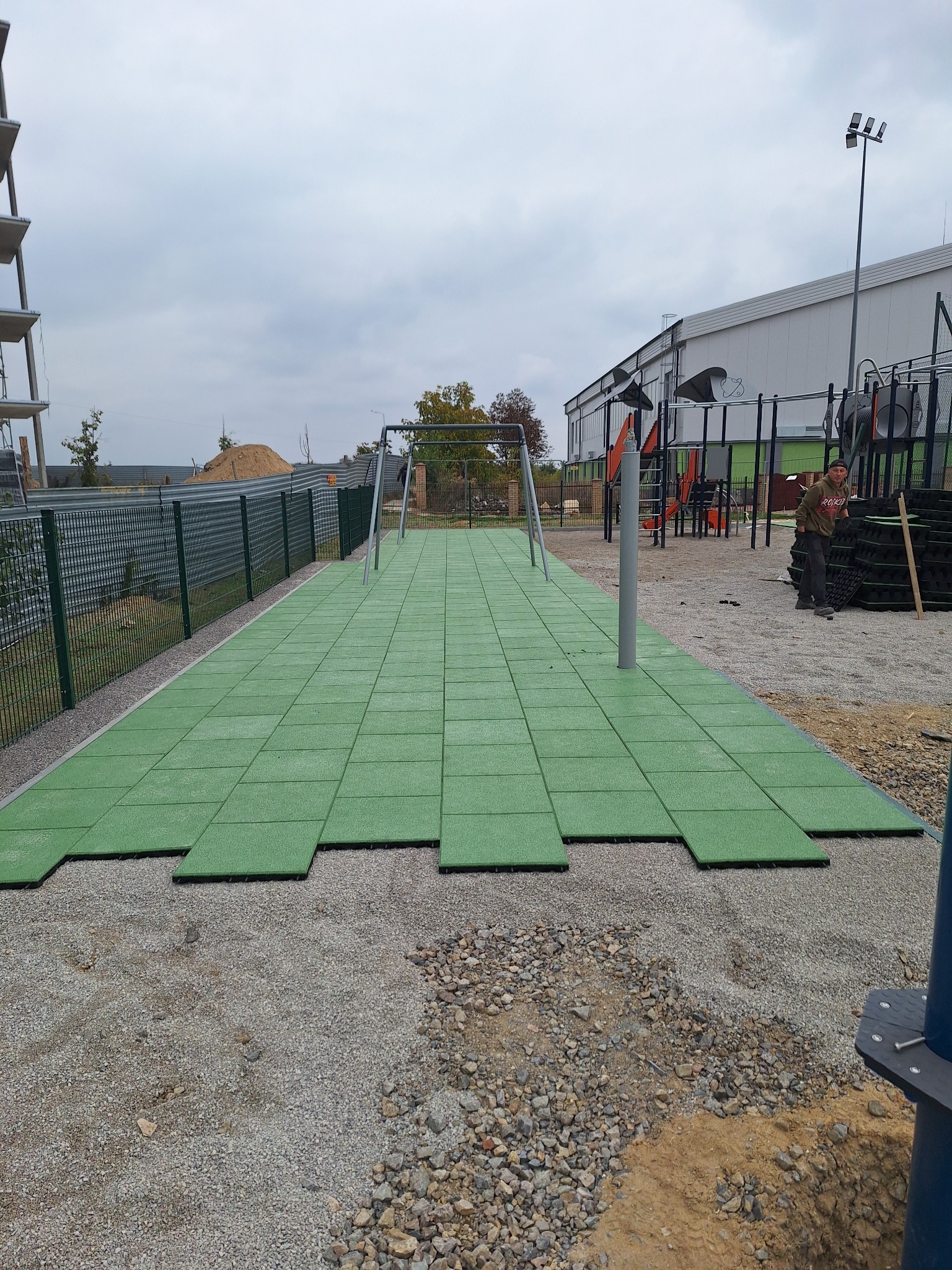 Montaż zielonej, gumowej nawierzchni na placu zabaw. Częściowo ułożone płyty, widoczny pracownik i elementy konstrukcyjne placu. Tło: budynek i pochmurne niebo.