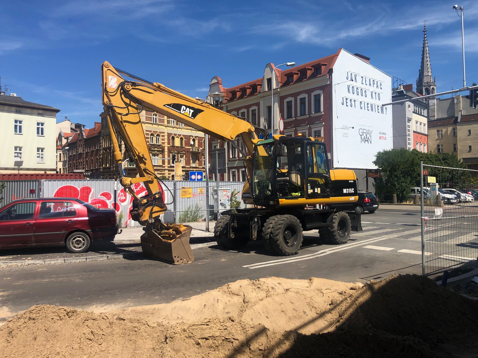 Żółta koparka kołowa CAT M315C na tle miejskiej zabudowy z reklamą filmu, obok zaparkowany czerwony samochód i pryzma piasku.