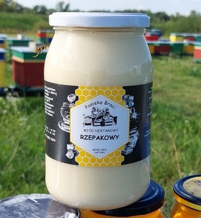 Słoik nektarowego miodu rzepakowego z etykietą Pasieka Braci na tle uli i latającej pszczoły.