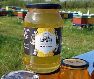 Słoik miodu akacjowego z czarną etykietą na tle uli i zielonej trawy, drugi słoik w dolnym rogu.