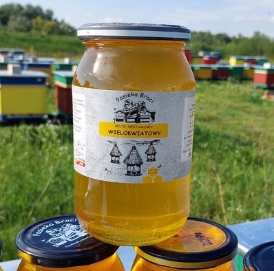 Słoik miodu wielokwiatowego z etykietą Pasieka Braci, na tle uli i zieleni, widoczny drugi słoik z pokrywką z logo pasieki.