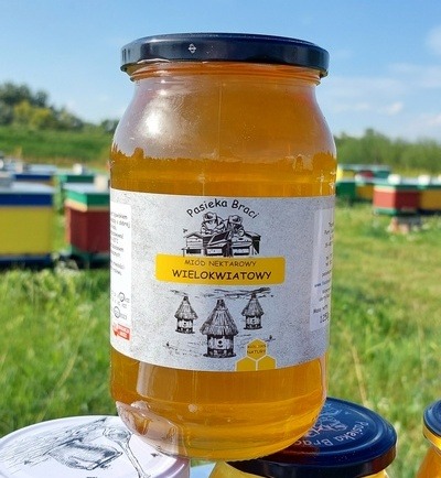 Słoik miodu wielokwiatowego z etykietą Pasieka Braci na tle uli i zieleni, widoczny fragment drugiego słoika w dolnej części kadru.