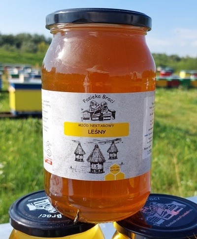 Słoik miodu leśnego z etykietą Pasieka Braci, ustawiony na pokrywkach innych słoików, z ulami w tle na zielonej łące.