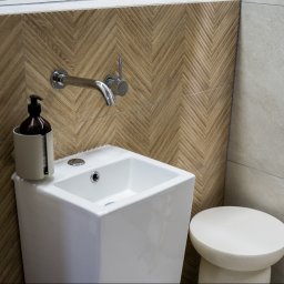 Araszkiewicz Nieruchomości na ARASZKIEWICZ NIERUCHOMOŚCI - Nowoczesna, biała umywalka z armaturą i dozownikiem mydła na tle ściany z drewnopodobnych płytek ułożonych w jodełkę. Minimalistyczny stołek obok.