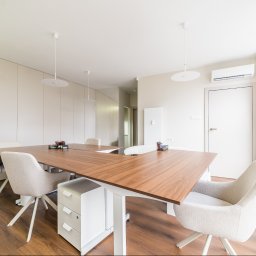 Araszkiewicz Nieruchomości na ARASZKIEWICZ NIERUCHOMOŚCI - Jasne biuro z drewnianym stołem, krzesłami na kółkach i białymi szafkami. Minimalistyczny design z okrągłymi lampami i drewnianą podłogą. Widok na okno z naklejką.